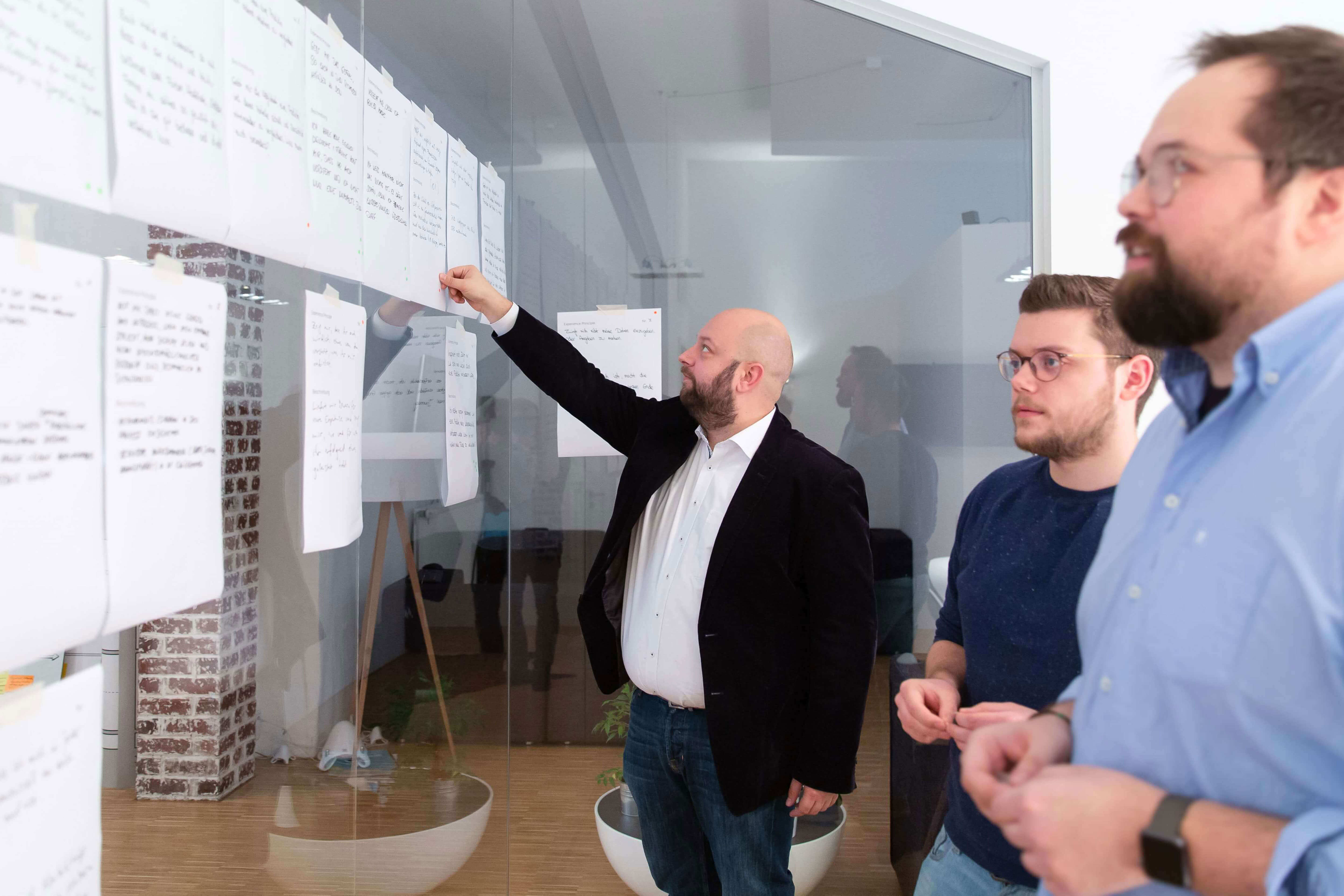 Team-Mitglieder von Me & Company stehen im Konferenzraum zusammen und betrachten Arbeitsergebnisse die an der Glasscheibe aufgehangen wurden