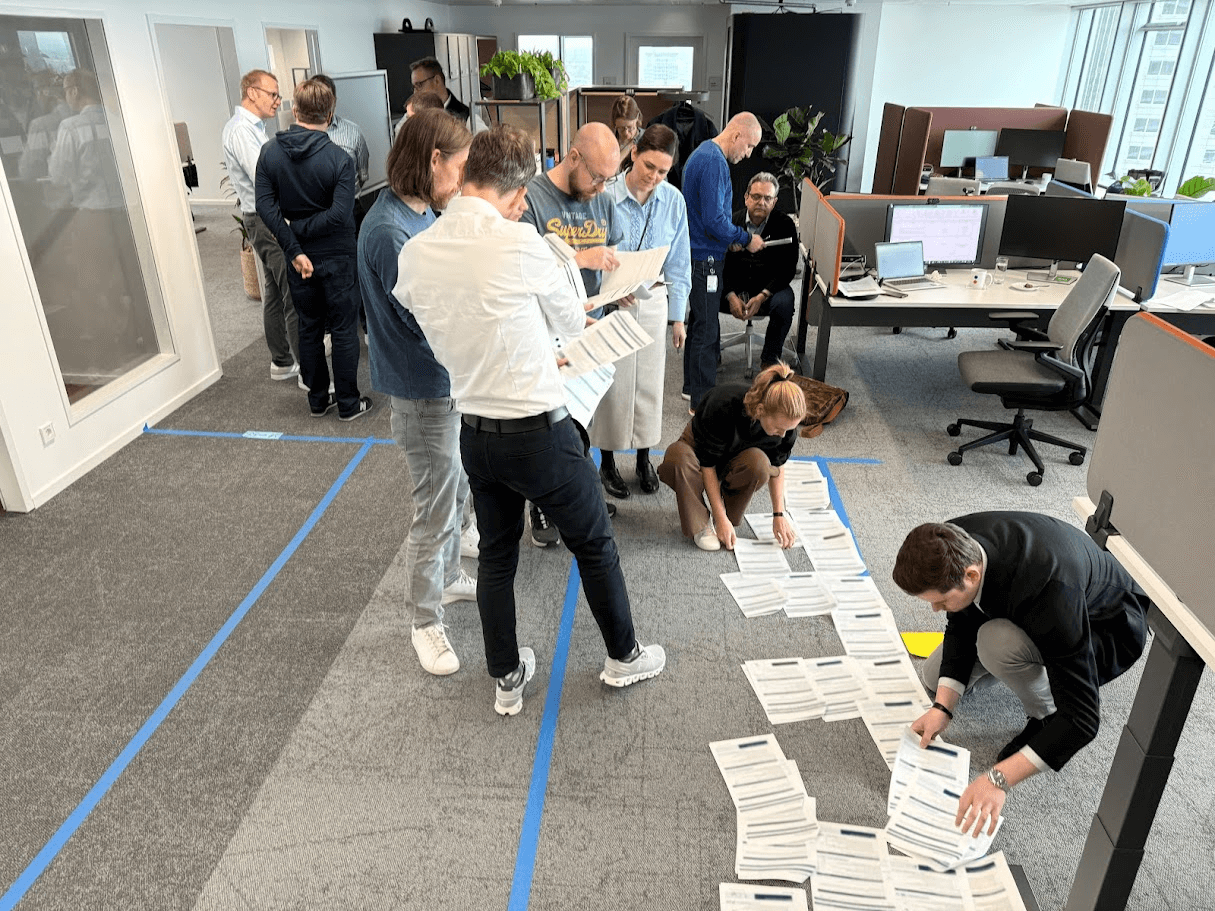 Ein Team steht in einem Raum, auf dem Zettel auf dem Boden verteilt sind, die von einigen Personen sortiert werden.